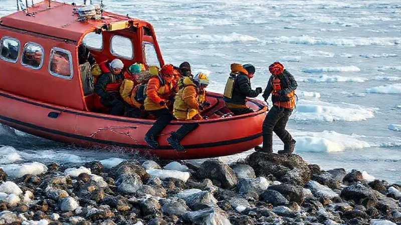 Photorealistic image of a group of well-dressed expedition passengers carefully disembarking a Zodiac onto a rocky, icy shore in the Arctic, guided by an experienced crew member. The passengers are wearing appropriate gear, demonstrating safe practices. Professional photography, 8K, cinematic lighting, sharp focus, depth of field, shot on a high-end DSLR.