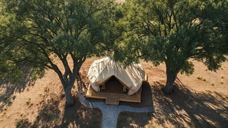 photorealistic, professional photography, 8K, cinematic lighting, sharp focus, depth of field, shot on a high-end DSLR, an aerial view of a meticulously planned glamping site, showing a luxury tent strategically placed under the partial shade of mature, healthy trees. The ground around the tent is well-drained, possibly with subtle gravel pathways, and the site is gently elevated, demonstrating intelligent environmental integration and protection.