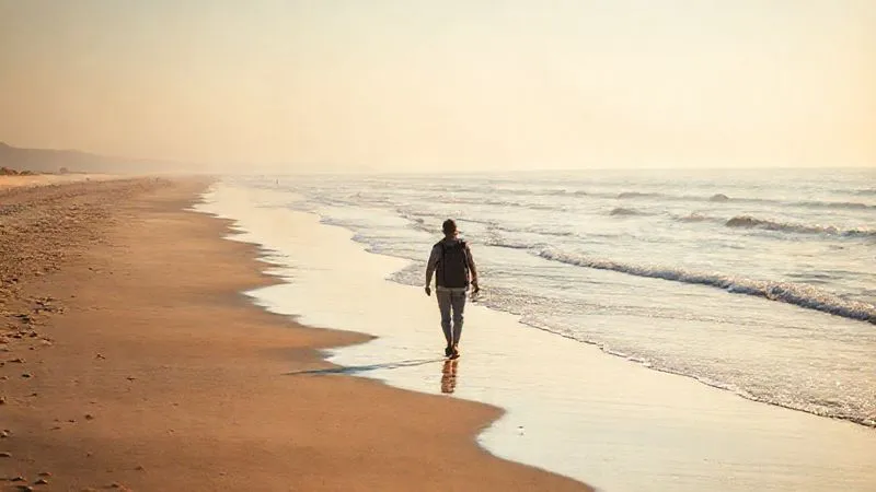 A photorealistic, professional photography image of a serene beach scene during shoulder season. The beach is sparsely populated, with soft, warm light of late afternoon. A single traveler walks peacefully along the shore, enjoying the tranquility. 8K, cinematic lighting, sharp focus on the traveler and the gentle waves, depth of field blurring the distant horizon, shot on a high-end DSLR.