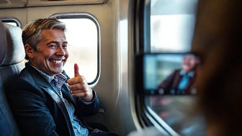 Photorealistic, professional photography, 8K, cinematic lighting, sharp focus, depth of field. A smiling, content client is seen on a train, looking out the window, giving a thumbs-up to their phone during a video call with their travel agent. The agent is partially visible on the phone screen, also smiling, conveying a sense of successful resolution and satisfaction.